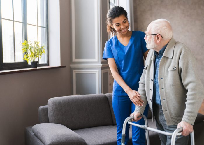 Portrait of an african young nurse helping old elderly disable man grandfather to walk using walker equipment in the bedroom. Senior patient of nursing home moving with walking frame and nurse support